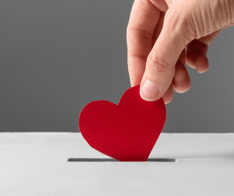 A hand holding a red paper heart is pushing it into the slot of a donation box