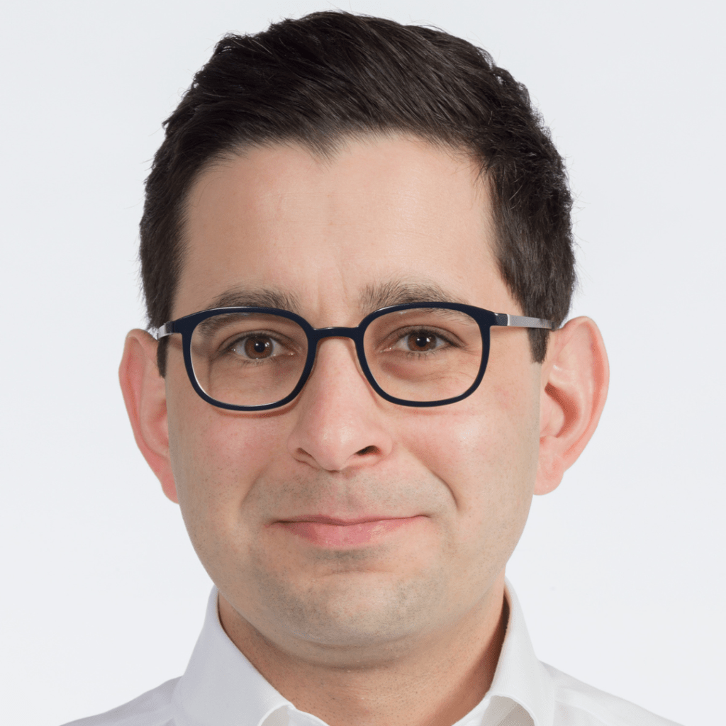 A picture of our new trustee Chris, he has black hair, black rimmed glasses. He is standing in front of a white background with a shirt shirt on smiling at the camera.