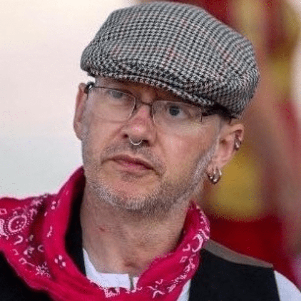 A picture of our new trustee John. He has a grey flatclap opn and glasses without frames. He is wearing a dark waistcoat and a pink bandana.