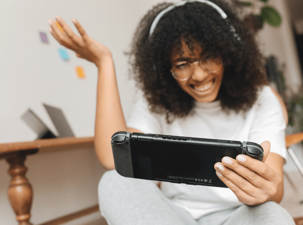A young girl with black afro hair and gold rimmed glasses is sitting on the floor. She has on a white Tshirt and grey jogging bottoms. On her head are headphones and in her hands is a games console. She looks excited and perplexed by the game she is playing.