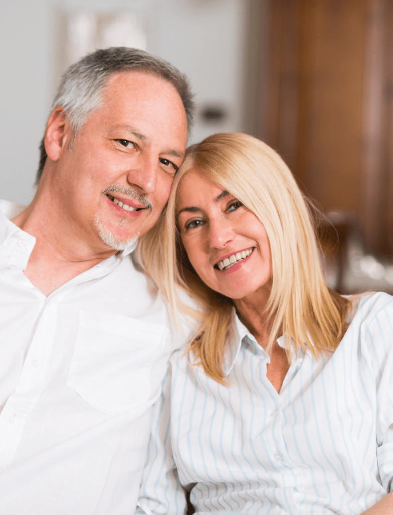 A couple in their fifties are sitting on a sofa looking content. They are resting their head on each other.
