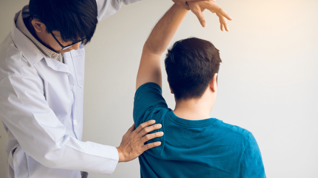 A man is sat in a chair with his arm raised as a medical person feels his shoulder movement and helps to hold the weight of his arm.