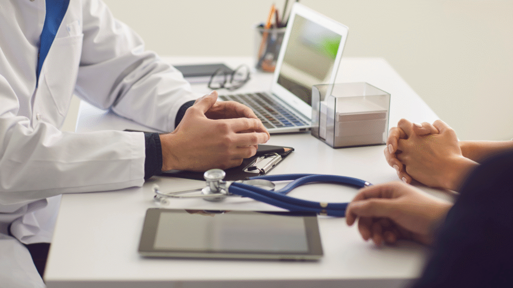 A man in a white coat is sat at a desk with his hands folded in front oh him. On his desk is a stethoscope, a blood pressure monitor and he is talking to patients about the risk of stroke being higher in different ethnicities