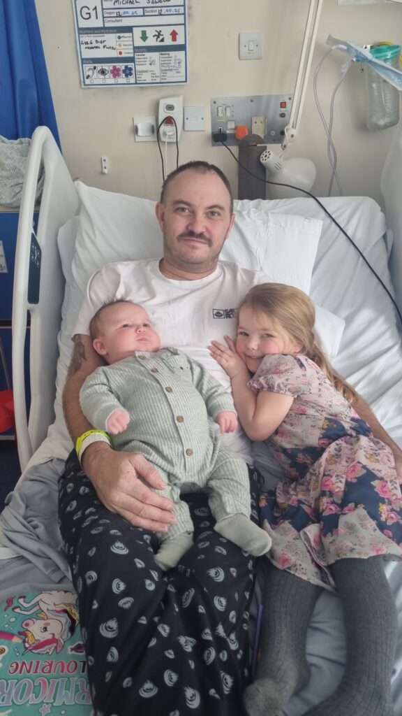 Mike who survived a haemorrhagic stroke in September 2025, aged 42 with two of his children in a hospital bed.